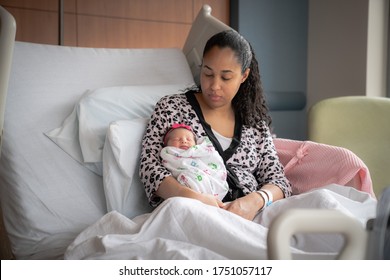 An African American Mixed Race Mother In A Pink Animal Print Bathrobe Sits On A Hospital Bed Looking At And Holding Her Newborn Baby Girl Swaddled In A Blanket.