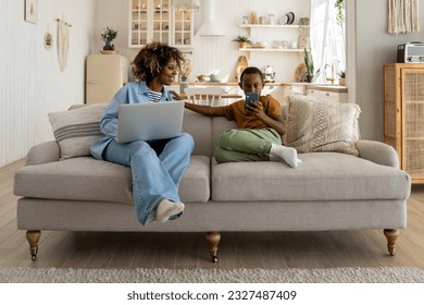 African american mom working on laptop and looking at smartphone in son's hands sitting on couch at home. Teen boy browsing in smartphone, watching videos. Working busy mom, child gadget addiction.  - Shutterstock ID 2327487409