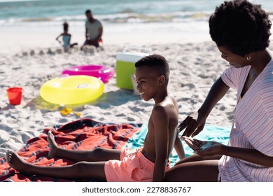 African american mother applying sunscreen on shirtless son's body while sitting on beach. Unaltered, family, together, picnic, protection, skincare, nature, vacation, enjoyment and summer concept. - Powered by Shutterstock