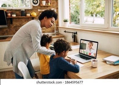 African American mother assisting her children who are learning online and having video call with their teacher over a laptop at home. 