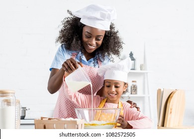 african american mother and daughter in chef hats pouring milk in bowl with ingredientes for dough 