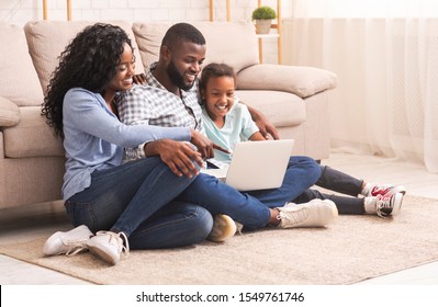 African american mother, father and daughter using laptop together watching family photos while sitting on floor at home.