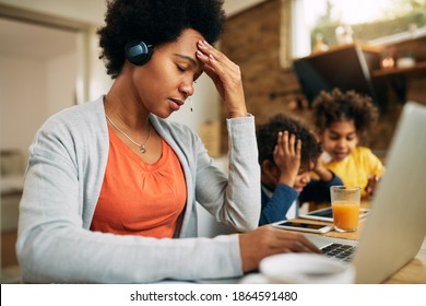 African American Mother Feeling Stressed Out While Working On Laptop At Home. Her Kids Are Surfing The Net On Touchpad Beside Her. 