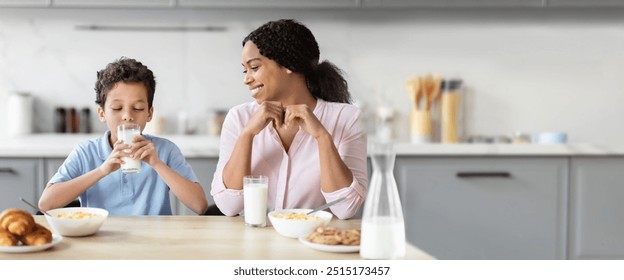 African American mother and her son share a joyful breakfast moment in their cozy kitchen. The boy drinks milk while surrounded by bowls of cereal and fresh pastries, creating a warm atmosphere. - Powered by Shutterstock