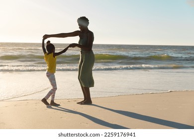 African american mother holding daughter's hands and dancing at shore against clear sky at sunset. Copy space, unaltered, family, summer, together, vacation, childhood, enjoyment and nature concept. - Powered by Shutterstock