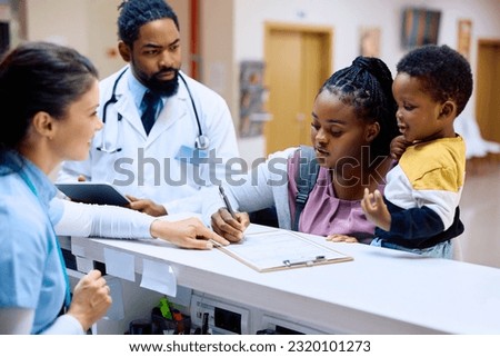 African American mother holding her small son while filling medical paperwork at reception desk in the hospital.