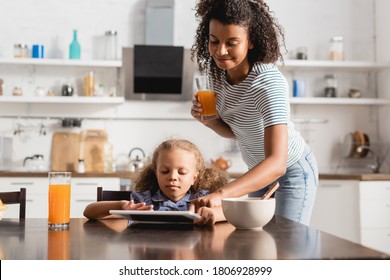 african american mother holding orange juice and touching digital tablet near concentrated child in kitchen - Powered by Shutterstock