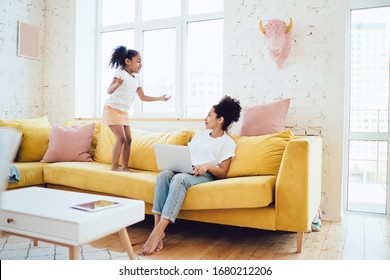 African American mother looking at daughter bouncing on yellow couch and drawing attention while working on laptop in cozy room at home - Powered by Shutterstock