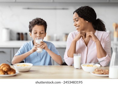 An african american mother observes her son drinking milk in a well-lit kitchen, capturing a family moment and the importance of a healthy breakfast - Powered by Shutterstock