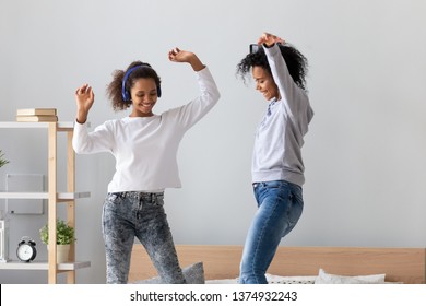 African American Mother With Phone In Hand And Teen Daughter Wearing Headphones Dancing To Favorite Music On Bed Together, Having Fun, Mum And Child Jumping, Enjoying Funny Activity At Home