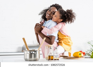 african american mother piggybacking daughter in apron on kitchen