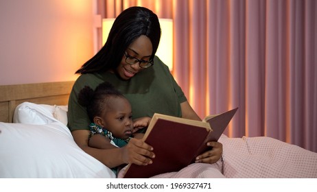 African American Mother Reading A Book To Her Little Daughter In Bed Before Going To Sleep At Home. Black Mom Story Telling To Girl In Bedroom