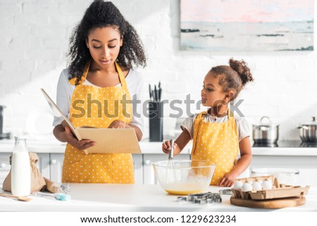 african american mother reading cookbook and daughter preparing dough in kitchen