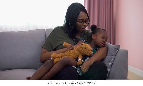 African American Mother Singing Lullaby For Little Daughter Going To Sleep On Sofa In Living Room . Black Mom Story Telling To Girl On Couch . Sleeping Child In Parent Embrace . Kid Hug Teddy Bear 