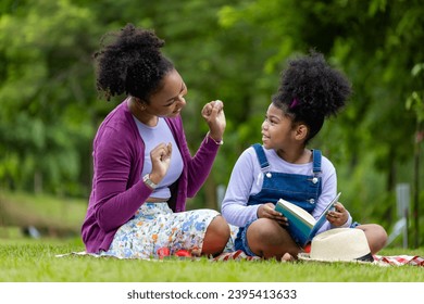 African American mother is teaching her young daughter to read using digital tablet while having a summer picnic in the public park for education and happiness - Powered by Shutterstock