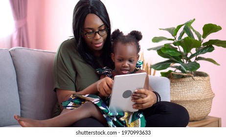 African American Mother Using Tablet With Her Little Daughter On Couch At Home . Black Girl Lying On Her Mom Learning Online At Home On Sofa Together . Quarantine Home School 