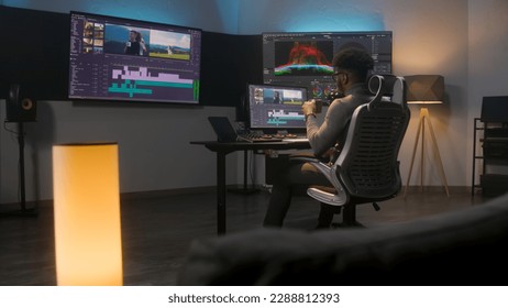 African American multimedia artist drinks coffee and makes color grading in editing program on computer. Process of video post production in modern studio. Multiple monitors with travel movie footage. - Shutterstock ID 2288812393