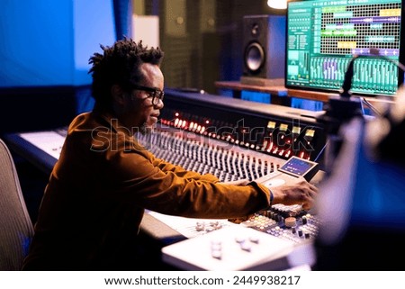 African american music producer composing tracks in control room, editing songs and adjusting volume settings with dashboard knobs. Audio engineer pressing faders and buttons.