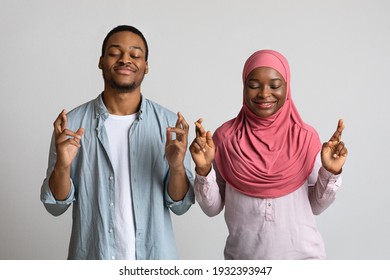 African American Muslim Couple With Eyes Closed And Fingers Crossed Standing Over Grey Studio Background. Superstitious Black Man And Woman In Hijab Praying Or Wishing For Luck And Protection