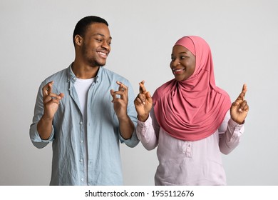 African American Muslim Couple With Fingers Crossed Looking At Each Other And Smiling Over Grey Studio Background. Superstitious Black Man And Woman In Hijab Praying Or Wishing For Luck And Protection