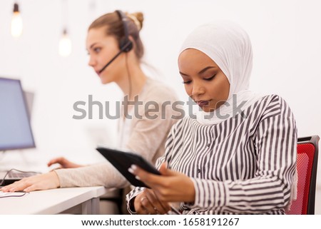 African american muslim girl with hijab working on a tablet in modern office. Caucasian girl colleague with headset in the background