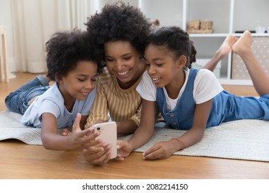 African American Nanny Teaching Kids To Use Online App On Smartphone. Happy Young Mother And Sibling Children Lying On Warm Floor, Making Video Call On Mobile Phone, Watching Content, Smiling