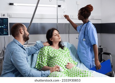 African american nurse checking pregnant woman iv drip, preparing for medical surgery in hospital ward. Patient with pregnancy lying in bed, discussing with husband about parenthood - Powered by Shutterstock