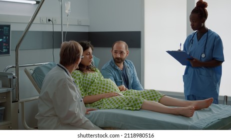 African American Nurse And Doctor Consulting Pregnant Woman Sitting In Hospital Ward. Multi Ethnic Medical Staff Talking To Expecting Mother And Father About Childbirth And Assistance