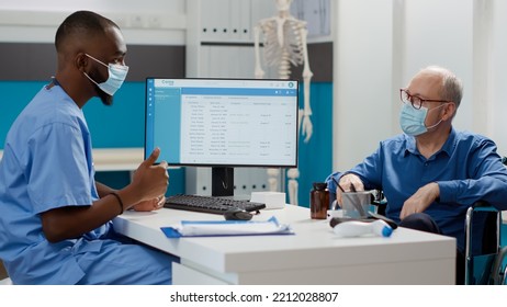 African American Nurse Doing Consultation With Elderly Man, Dealing With Physical Impairment In Wheelchair. Having Checkup Visit Appointment With Patient During Covid 19 Pandemic.