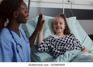 African American Nurse Doing High Five Gesture With Sick Girl Resting In Hospital Pediatric Ward Patient Bed. Childcare Healthcare Facility Staff High Fiving Ill Kid Under Medical Treatment.