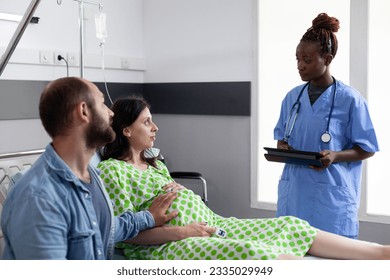 African american nurse explaining labor process to pregnant woman, preparing couple for child delivery in hospital ward. Patient with pregnancy lying in bed while husband comforting about motherhood - Powered by Shutterstock