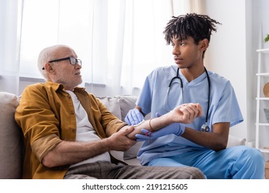 African American Nurse In Latex Gloves Holding Cotton Pad On Hand Of Senior Patient At Home