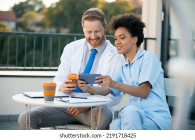 African American Nurse And Male Doctor Using Mobile Phone While Having Coffee Break On A Terrace At The Hospital.