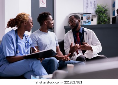 African American Nurse And Physician Talking To Person, Doing Consultation With Young Adult In Waiting Room At Hospital Lobby. Medical Team Helping Man With Treatment And Medcine In Waiting Area.