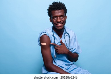 African American Nurse Pointing To Vaccine Shot Bandage While Looking At Camera And Smiling. Black Medical Assistant Showing Adhesive Plaster After Getting Vaccinated Against Coronavirus