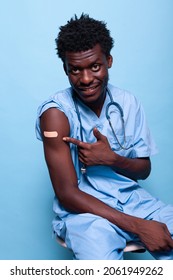 African American Nurse Pointing To Vaccine Shot Bandage While Looking At Camera In Studio. Black Healthcare Assistant Showing Adhesive Plaster After Getting Vaccinated Against Coronavirus