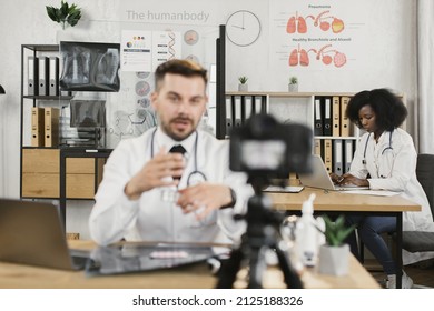 African American Nurse Typing On Modern Laptop While Qualified Caucasian Doctor Recording Video On Camera About Pills. Concept Of People, Technology And Health Care.