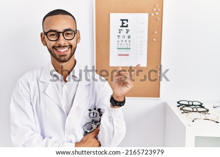 African american optician man standing by eyesight test with a big smile on face, pointing with hand finger to the side looking at the camera. 