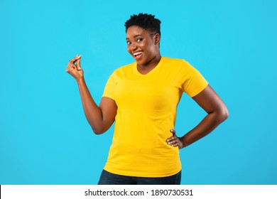 African American Oversized Woman Gesturing Clicking Fingers Standing Posing On Blue Background In Studio, Smiling To Camera. Finger Snap Gesture Concept