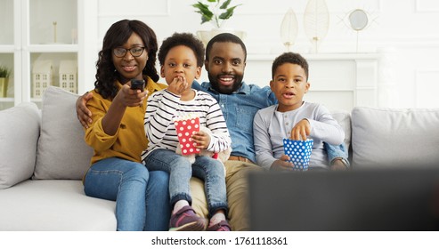 African American Parents With Little Kids Watching Funny Movies Or Tv Show, Eating Popcorn, Resting On Sofa At Home. 