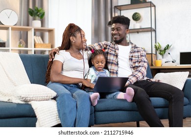 African American Parents Looking On Each Other With Smile On Face While Cute Little Daughter Sitting Among Them With Laptop On Knees. Family Tie At Home With Modern Technology.