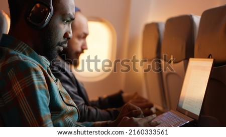 African american passenger in airplane seat flying on work trip, using laptop on flight. Travelling with commercial airline during sunset, wearing headphones on aviation jet. Handheld shot.