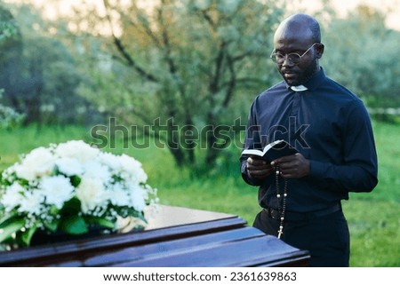 African American pastor with open Holy Bible carrying out funeral service at graveyard while standing by wooden coffin with white flowers on top
