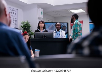 African American Patient Registering For Doctor Appointment At Private Clinic Front Desk Talking With Medical Doctor And Receptionist. Diverse People Having Conversation In Waiting Room.