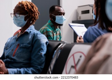 African American Patient Wearing Face Mask Talking To Assistant In Waiting Area Of Hospital Reception Lobby. Man Doing Healthcare Consultation With Medical Worker, Nurse And Person At Medical Facility