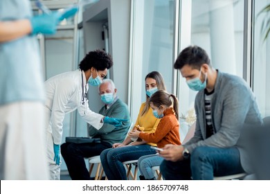 African American Pediatrician Communicating With A Little Girl In Waiting Room At Medical Clinic. They Are Wearing Face Masks Due To COVID-19 Pandemic.