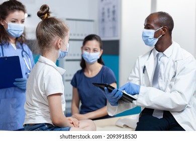 African American Pediatrician Doctor With Protection Face Mask Against Coronavirus Explaining Medical Treatment To Family During Clinical Appointment In Hospital Office. Health Care Service