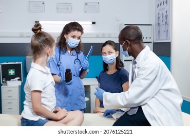 African American Pediatrician Doctor With Protective Face Mask Against Coronavirus Discussing Medication Treatment With Family During Clinical Appointment In Hospital Office. Health Care Service