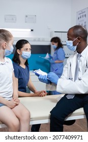 African American Pediatrician Doctor Writing Disease Symptoms On Clipboard Discussing Healthcare Treatment With Family During Clinical Examination In Hospital Office. Medical Team With Face Mask