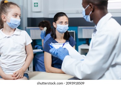 African American Pediatrician Doctor Writing Sickness Symptoms On Clipboard Discussing Healthcare Treatment With Family During Clinical Appointment In Hospital Office. Medical Team With Face Mask